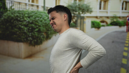 Young hispanic man in gray shirt experiencing back pain stands on a city street outdoors with a pained expression surrounded by urban greenery and buildings.