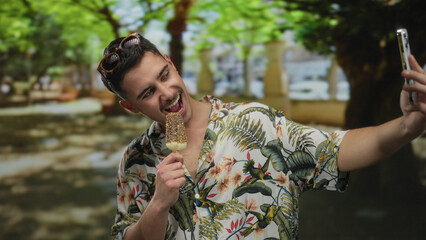 Young man enjoying ice cream while taking a selfie in a lush park setting with vibrant flora,...