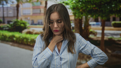 Fototapeta premium Young hispanic woman in blue shirt wearing glasses holds throat with hand on street near buildings; panic suffering.