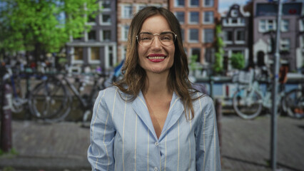 Fototapeta premium Smiling woman wearing glasses raising hand in ok sign on a busy city street lined with parked bicycles and leafy trees; approval.