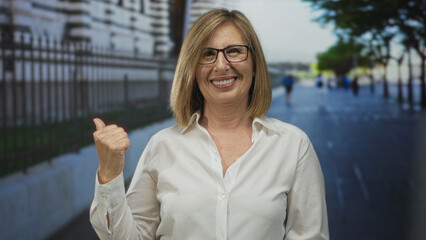 Fototapeta premium Smiling woman wearing glasses and white shirt shows thumb up gesture on city street pavement; optimism.