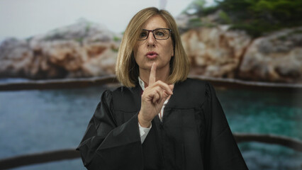 Middle age caucasian woman judge wearing black robe finger to lips on street by rocky seaside...
