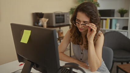 Woman adjusts glasses while viewing computer monitor on white desk in building; concentration...