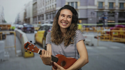 Young hispanic woman smiling while hugging a ukulele with her hands on a busy city street lined with taxis and construction barriers; joy.