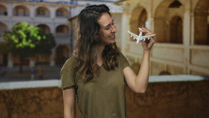 Woman smiles and holds model plane in hand under ancient stone arches on old town street; wanderlust.