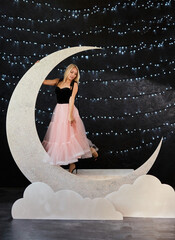 A beautiful blonde woman in a black top and a voluminous pink tulle skirt poses in a large glitter crescent moon prop. The backdrop is a dark studio illuminated by fairy lights and white cloud props.
