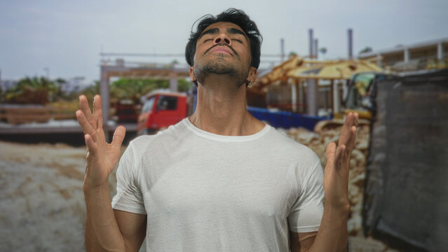 Young man tilting head back with closed eyes and bare arms raised while clenching fists at sunlit building site; relief.