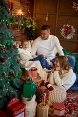 A happy family mother, father, and young son sitting close together in a cozy, festive living room and decorating a Christmas tree.