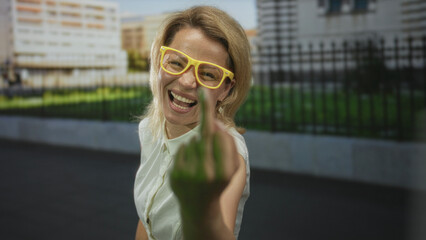 Woman wearing bright yellow glasses raises middle finger on street in front of building fence;...