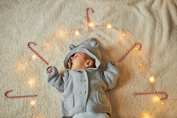 A newborn baby in a gray knit hooded outfit with bear ears is peacefully sleeping, surrounded by glowing fairy lights and candy canes on a soft, textured blanket. Overhead shot