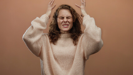 Woman expressing frustration in a beige sweater against a brown wall backdrop, isolated for...