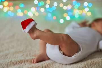 Close-up shot of a newborn baby lying on its back with its tiny feet in the air, one of which has a miniature red Santa hat perched on the toes.