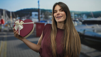 Young hispanic woman holds heart-shaped gift box near port with boats and sea in the background,...