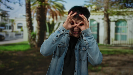 Young man in denim jacket making glasses gesture with fingers in a park setting, showcasing playful outdoor vibe with trees and architecture in background.