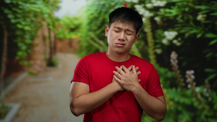 Fototapeta premium Young man in red shirt holding chest in park setting with green lush background, displaying emotion and introspection outdoor.