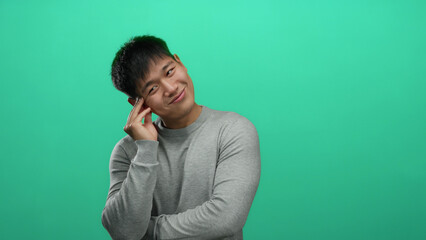 Young man in grey sweater with hand on face over isolated green background exuding contemplation...