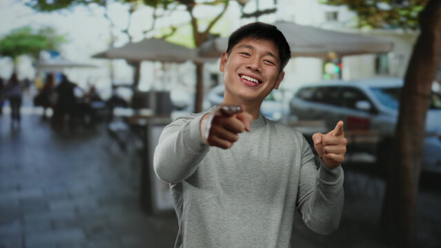 Young asian man smiling cheerfully while gesturing on a sunny restaurant terrace with blurred outdoor seating and people in the background. - Powered by Adobe