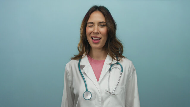 Hispanic female doctor in white coat with stethoscope winks playfully against isolated blue background embodying professionalism and warmth.