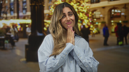 Young blonde woman clasped hands in pleading prayer gesture at holiday market; hope apology longing.