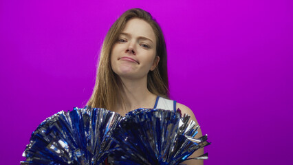 Young cheerleader woman smirks while holding metallic blue pompom in studio; confidence empowerment.