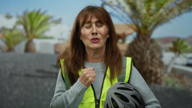 Senior hispanic woman standing outdoors on a city street wearing a safety vest and holding a helmet, with palm trees in the background.