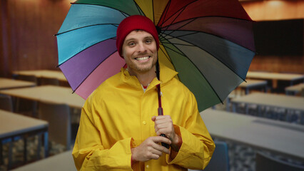 Young man in yellow raincoat smiling under colorful umbrella in an empty classroom