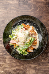 Grilled chicken Caesar salad with romaine lettuce, cherry tomatoes, croutons, Parmesan cheese, and creamy dressing served on a black plate over rustic wooden background.