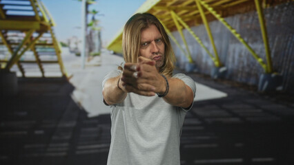 Blond man with long hair forming finger gun with hand while pursing lips on urban street;...