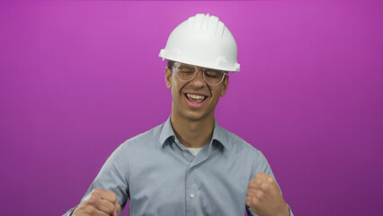 Young hispanic engineer in hardhat helmet and eyeglasses clenches both fists in purple studio; excitement.