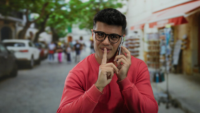 Young man smiling while talking on smartphone outdoors, standing on a vibrant city street with trees and blurred people in the background, wearing glasses and a red sweater.