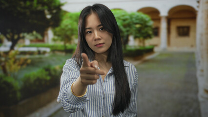 Young chinese woman pointing outdoors with a determined expression against a blurred garden and...