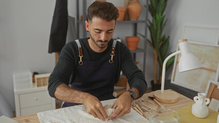 Hispanic man shapes clay sculpture with bare hands on a wooden table in a bright studio; creative focus.