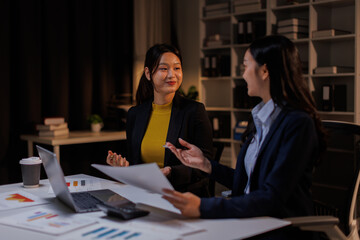 In a dimly lit office, business asian women collaborate late into the night. This image captures the intensity and focus of teamwork necessary for project success.

