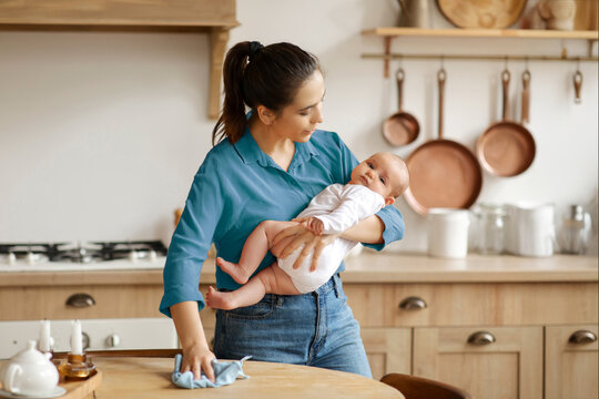 Mother holding baby on hand and cleaning table in kitchen with cloth, doing house chores, housework with child. Hygiene and multitasking concept