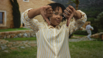 Young african american woman lifts both thumbs down gesture in front of old stone building;...