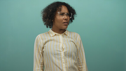 Woman wearing a striped shirt and glasses looking aside with a neutral expression in a teal studio;...
