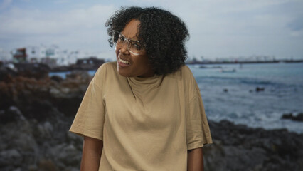 Woman grimaces with bare face and translucent glasses at ocean waves on rocky seaside coast;...