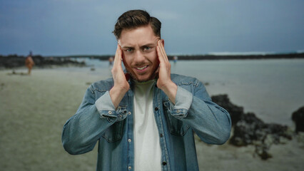 Young man with beard feeling stressed at the seaside, standing on a beach with ocean waves in the...