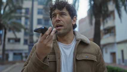 Hispanic man speaking on smartphone outdoors on a city street lined with palm trees, exuding...
