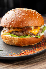 Close-up of gourmet hamburger with brioche bun, beef patty, cheddar cheese, lettuce, caramelized onions, and sauce on dark plate, sprinkled with chili flakes.