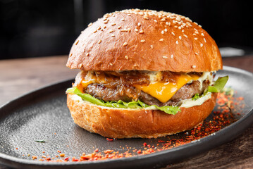 Close-up of gourmet hamburger with brioche bun, beef patty, cheddar cheese, lettuce, caramelized onions, and sauce on dark plate, sprinkled with chili flakes.