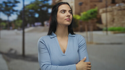 Fototapeta premium Young hispanic woman standing thoughtful on a sunny outdoor street wearing blue with a background of trees and buildings reflecting serene city life.