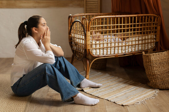 Young woman sitting tired and depressed by the crib while baby sleeping. Postpartum depression concept - Powered by Adobe