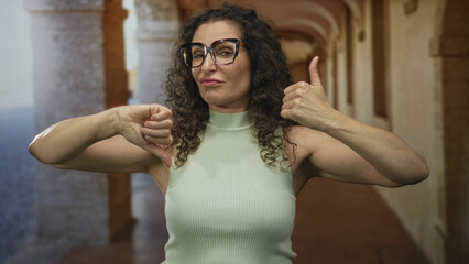Young woman shows both thumbs up and thumbs down gesture in a narrow sunlit street in old town;...
