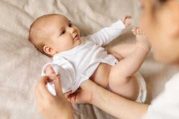 Mother dressing up, changing bodysuit of her cute little baby after waking up, kid lying on bed....