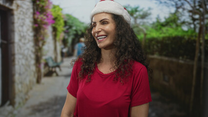 Hispanic woman in red shirt smiling with open mouth and visible teeth under santa hat on narrow...