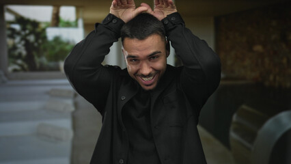 Man with bare hands making bunny ears above his smiling face in an indoor building space; playfulness.