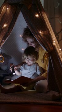 Mother and child reading bedtime story in cozy tent illuminated by fairy lights vertical slow motion - warm family evening atmosphere representing gentle motherhood and peaceful childhood moments