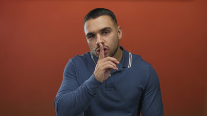Young hispanic man presses finger to lips for silence gesture in a red studio setting; silence...