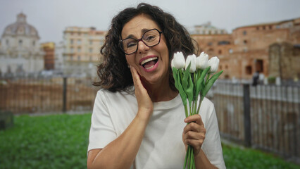 Woman holding bouquet of white flowers in front of roman ruins outdoors with joyful expression in vibrant daylight setting.